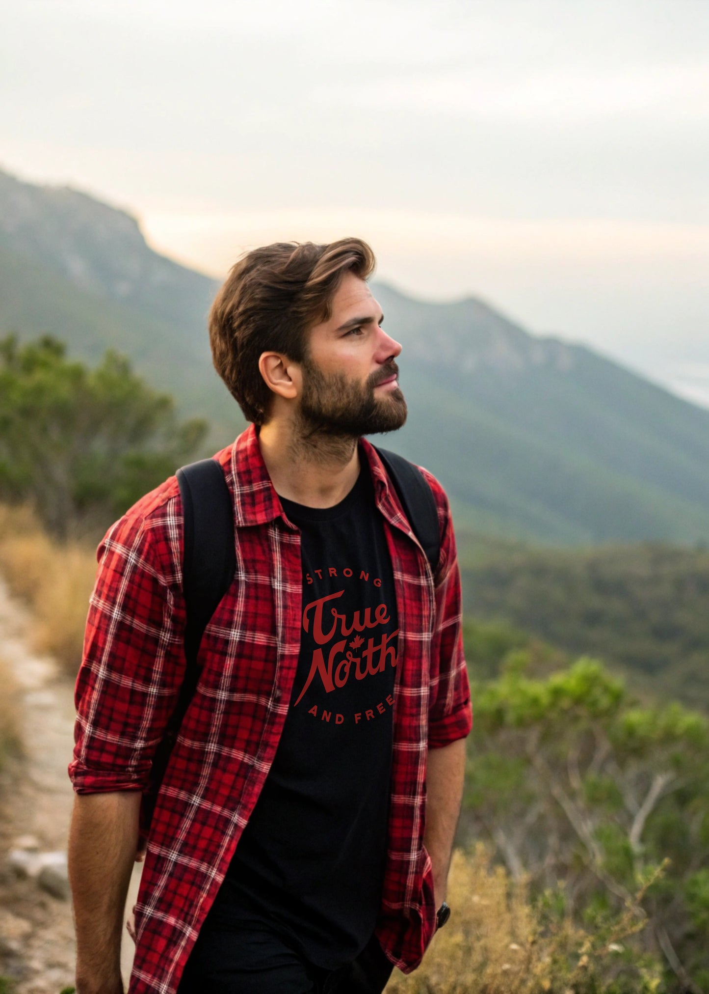 Man in a red plaid shirt and black t-shirt with 'True North Strong and Free' graphic, standing in a mountainous landscape.