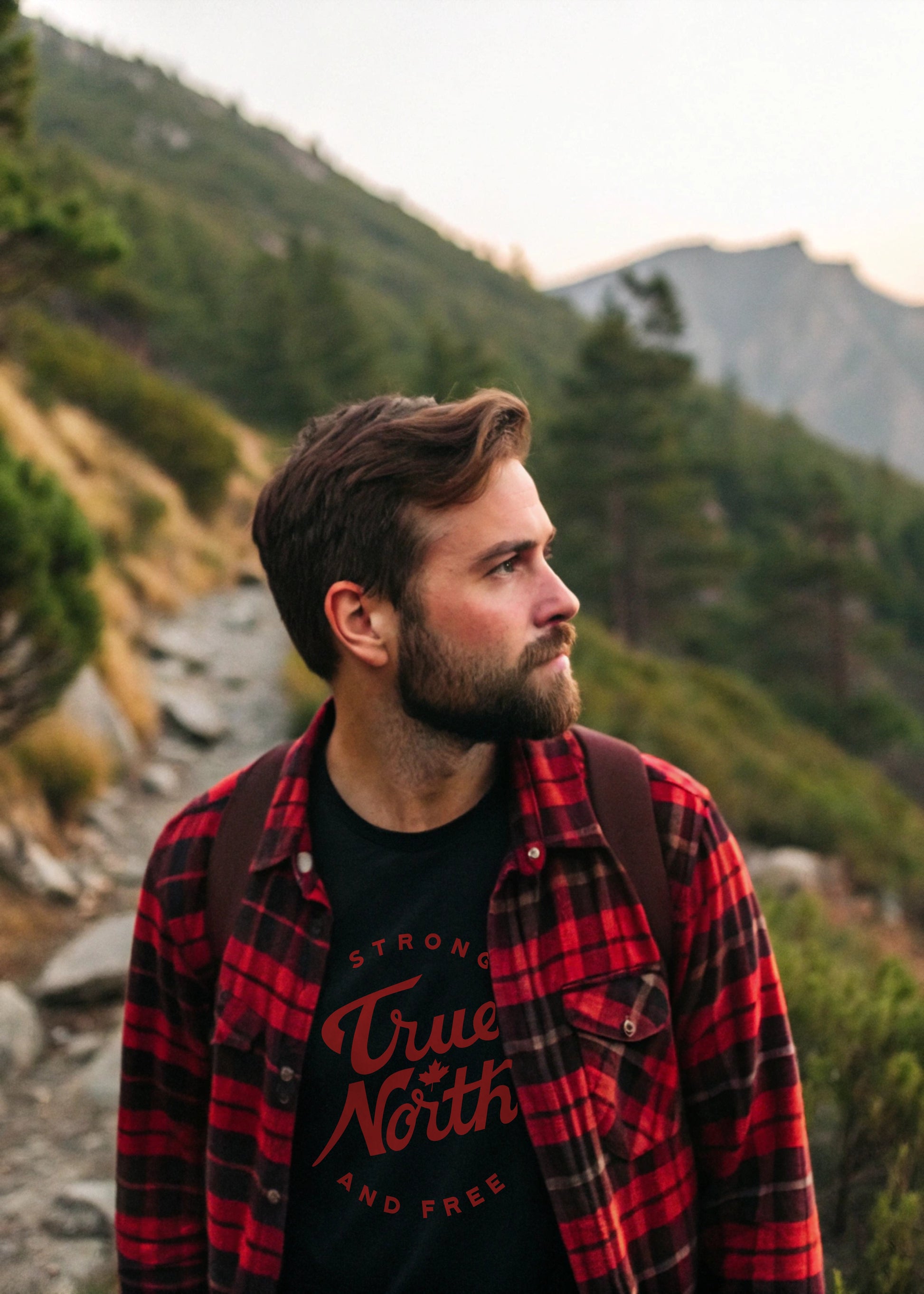 Man in a red plaid shirt and black t-shirt with 'True North Strong and Free' graphic, standing in a mountainous landscape.
