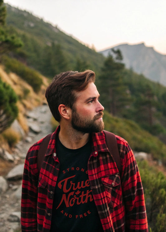 Man in a red plaid shirt and black t-shirt with 'True North Strong and Free' graphic, standing in a mountainous landscape.