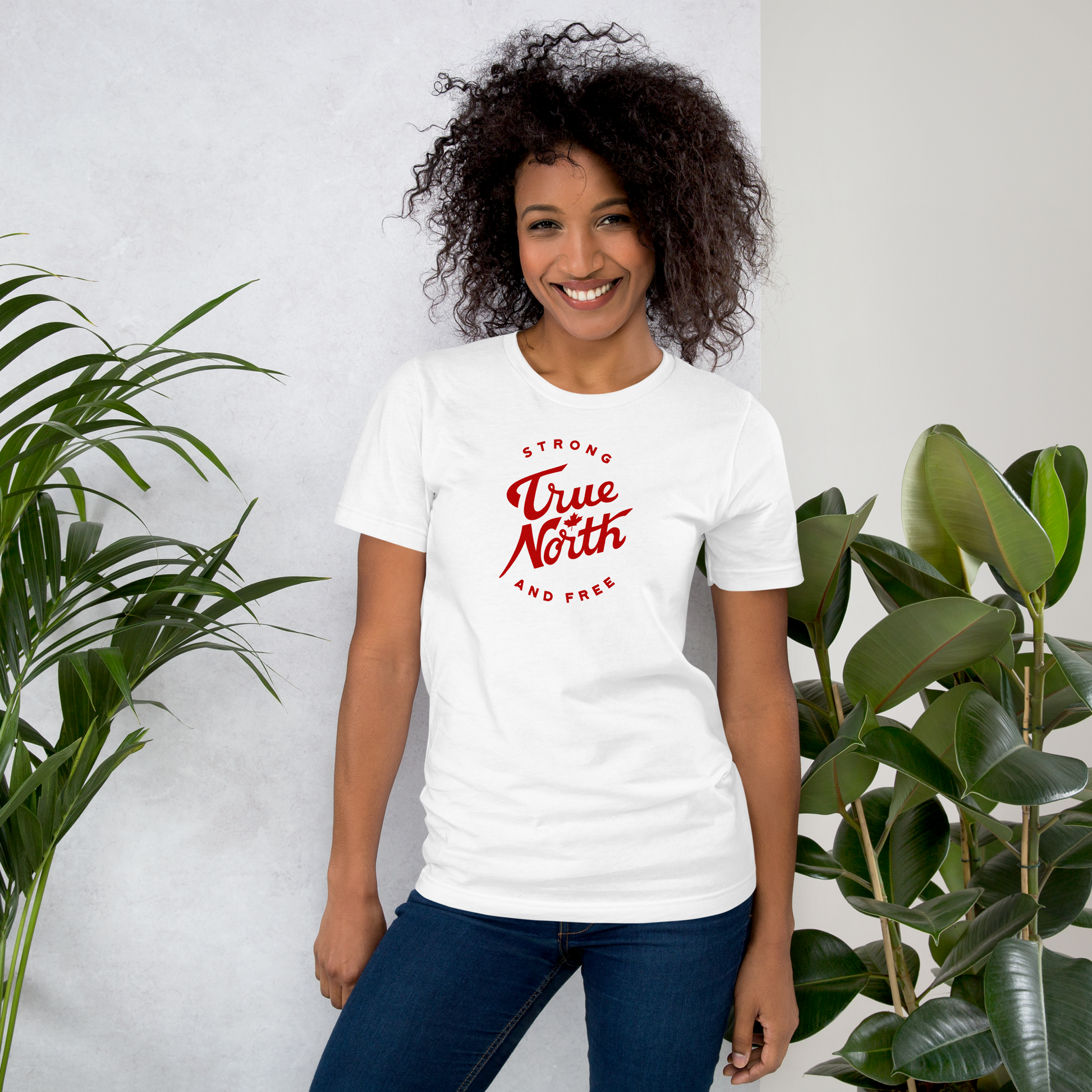 Woman wearing a white t-shirt with red text with 'True North Strong and Free' text standing between two plants against a light gray background. 